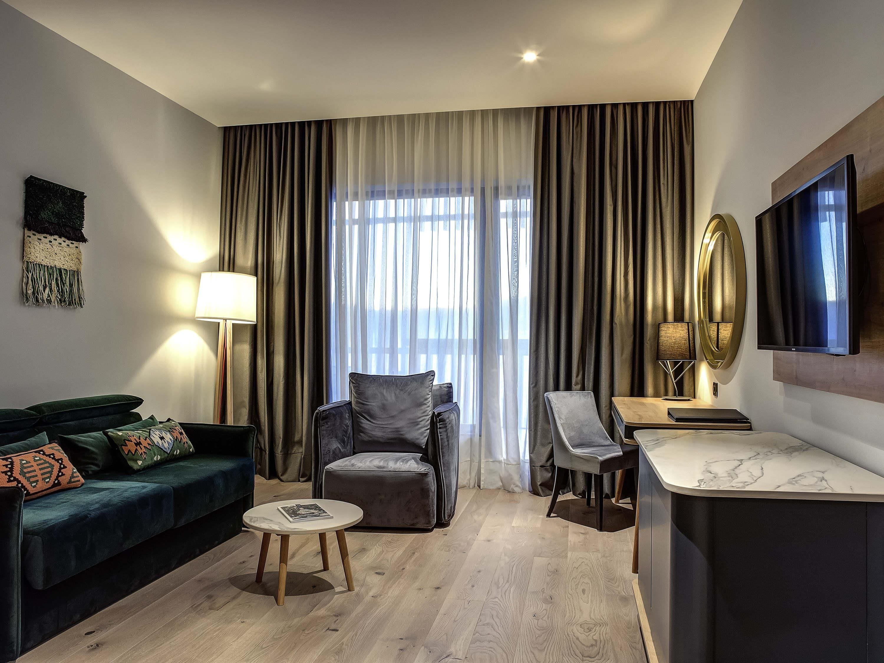 A modern hotel room with a dark green sofa, a small round table, a grey armchair, a wooden desk, and a large window with curtains.
