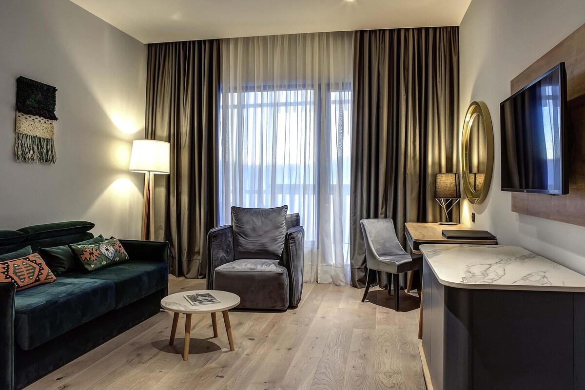A modern hotel room with a dark green sofa, a small round table, a grey armchair, a wooden desk, and a large window with curtains.