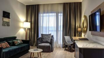 A modern hotel room with a dark green sofa, a small round table, a grey armchair, a wooden desk, and a large window with curtains.