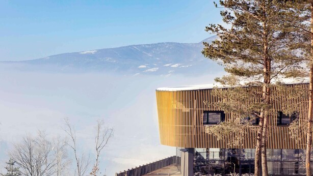 Ein modernes Gebäude mit Holzfassade und großen Fenstern, umgeben von schneebedeckten Bäumen und einer Bergkulisse.