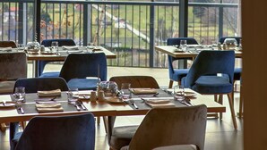 Ein Restaurant mit großen Fenstern, das einen Blick auf Berge und eine ferne Stadt bietet. Tische sind mit Geschirr gedeckt und die Stühle blau gepolstert.