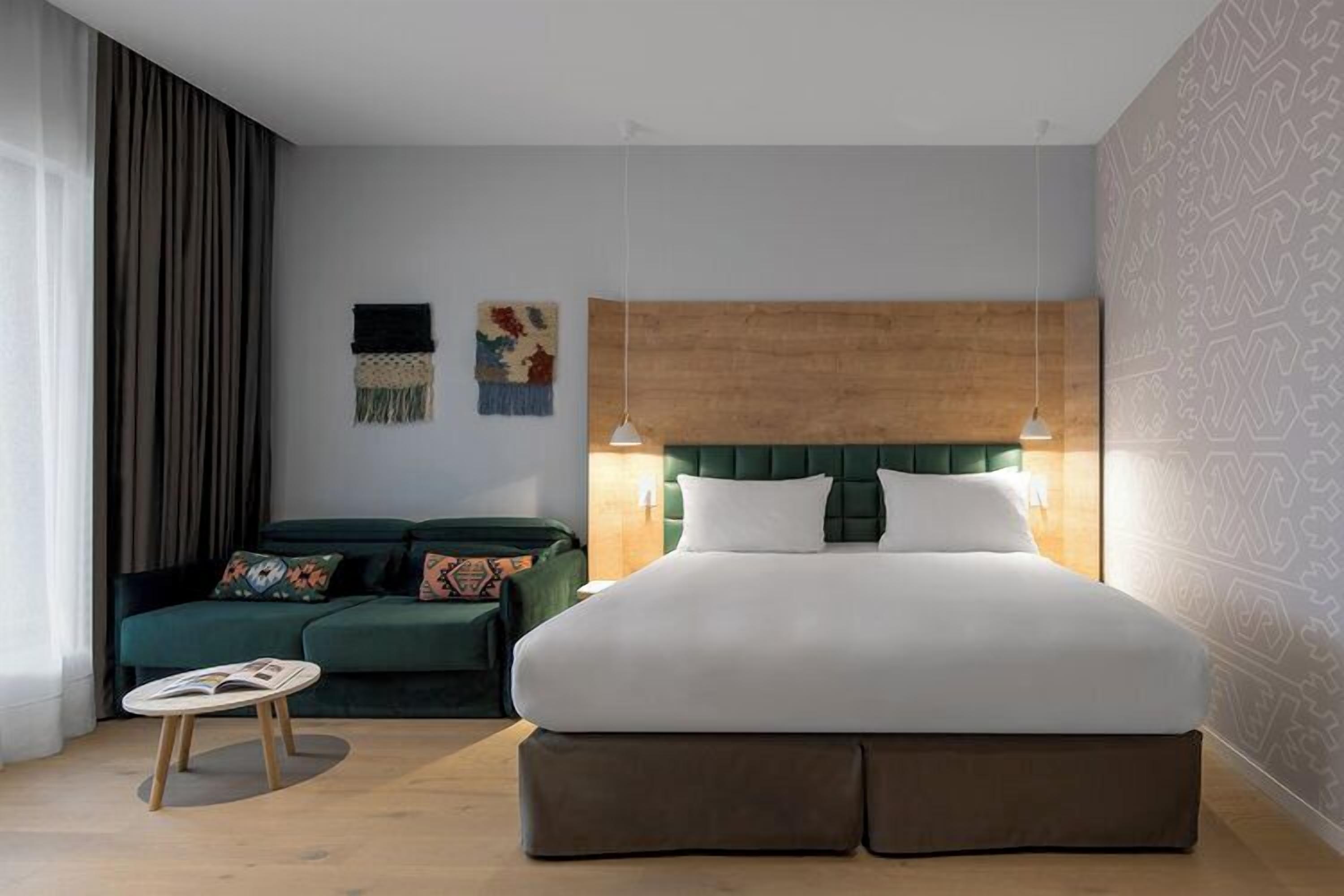 Une chambre d’hôtel moderne avec un grand lit, un canapé vert, une petite table ronde et un mur décoré de motifs.