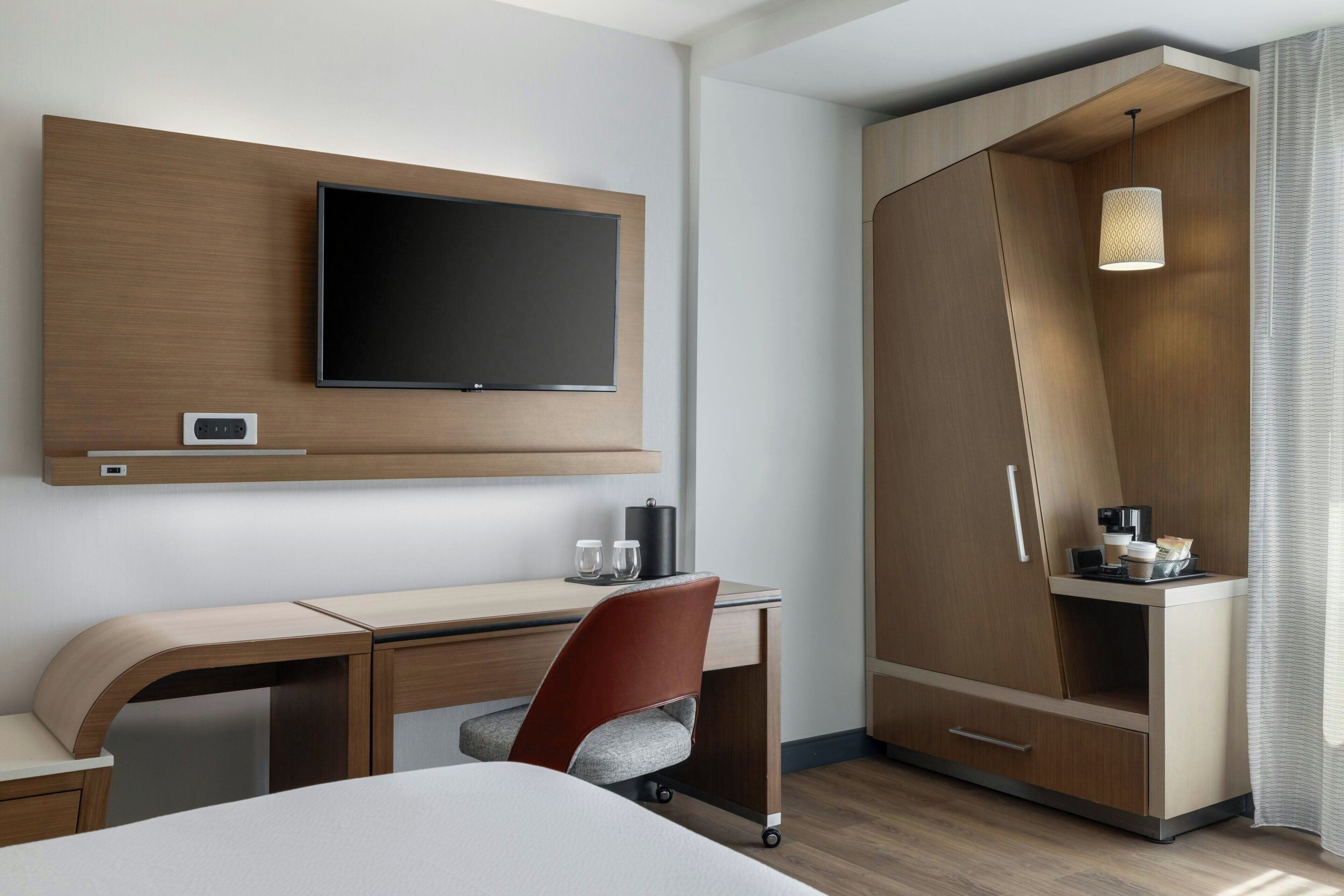 Una habitación de hotel moderna con televisor de pantalla plana, escritorio, silla, armario e iluminación colgante.