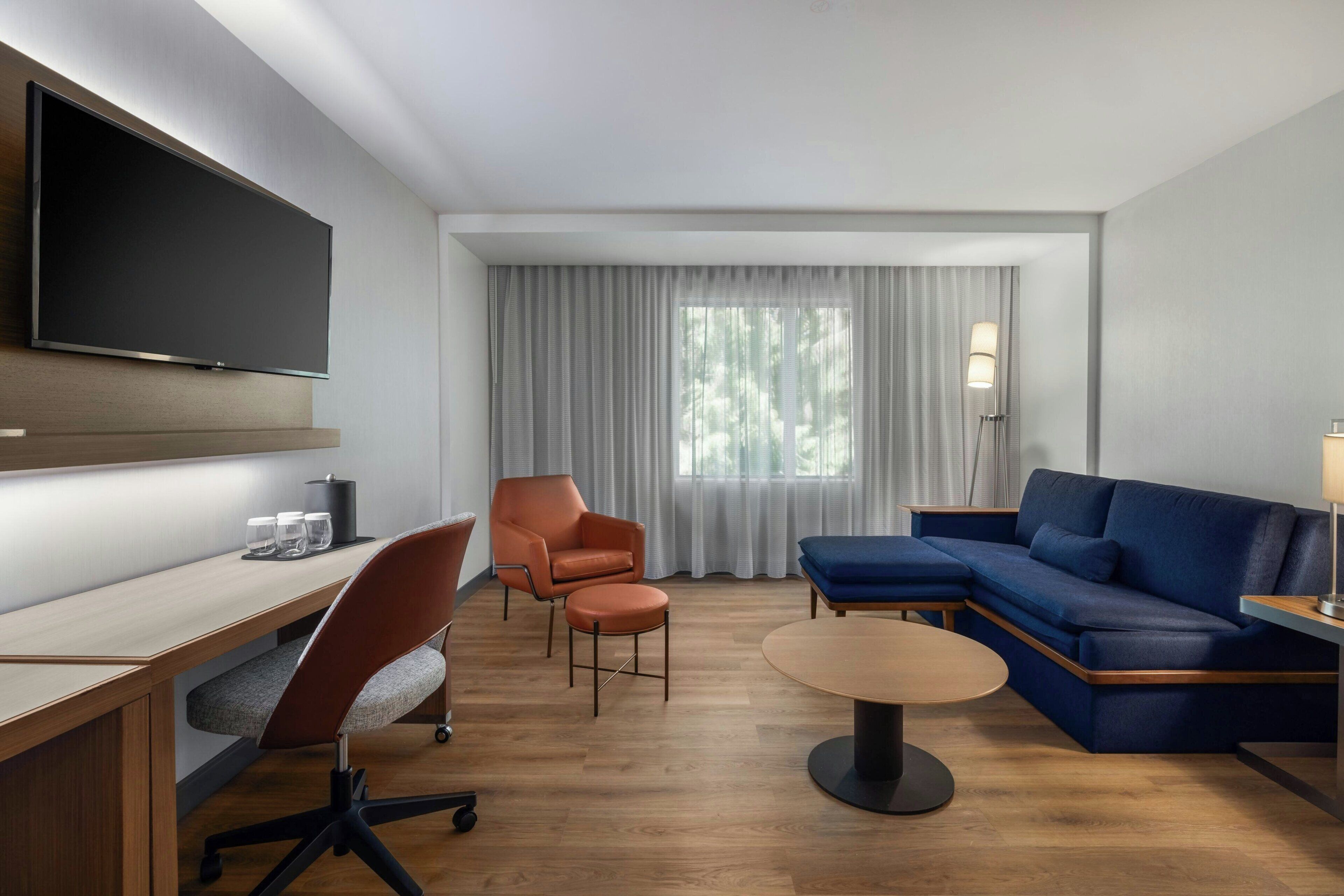 Habitación de hotel moderna con televisión de pantalla plana, escritorio con vasos, sofá azul y una ventana grande con cortinas.