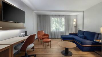 Habitación de hotel moderna con televisión de pantalla plana, escritorio con vasos, sofá azul y una ventana grande con cortinas.