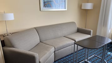 Et hotelværelse med en grå sofa, et lille bord, to lamper og et mønstret tæppe.