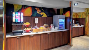 Un mostrador de buffet con una exhibición de frutas, un dispensador de bebidas y un refrigerador.