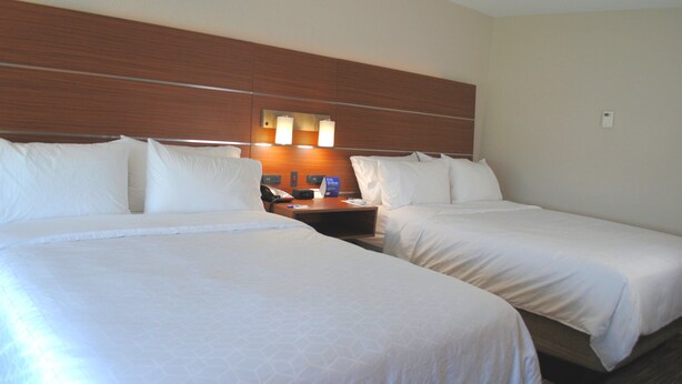 A hotel room with two beds, a wooden headboard, a nightstand with a phone and lamp, and a wall-mounted light fixture.