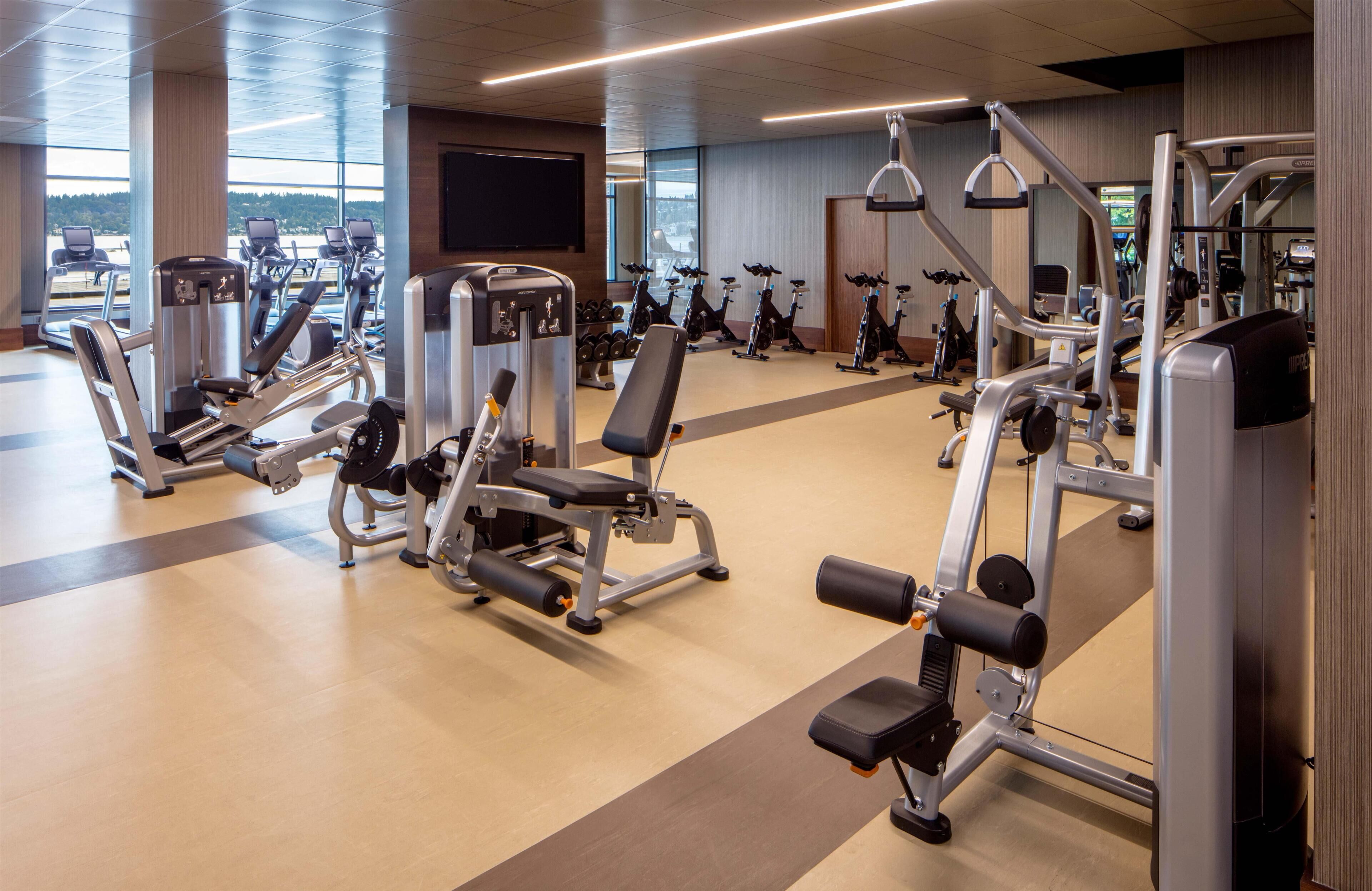 A gym with various exercise machines, a large TV screen, and windows offering a view of the outdoors.