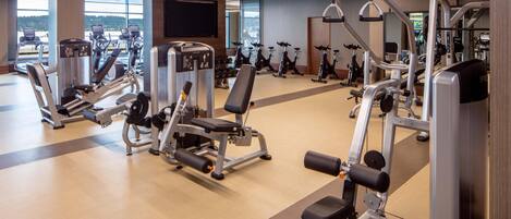A gym with various exercise machines, a large TV screen, and windows offering a view of the outdoors.
