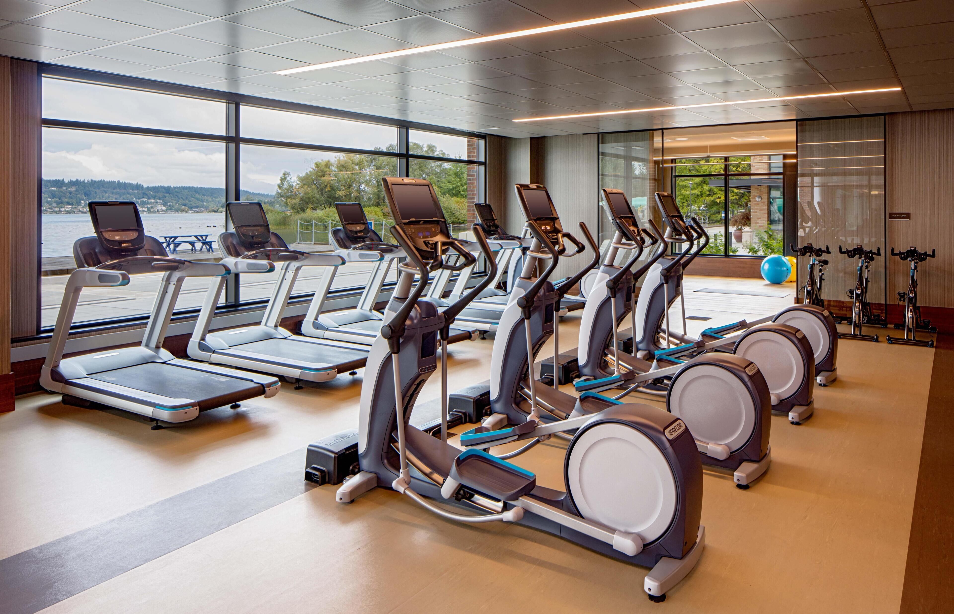 Une salle de sport équipée de plusieurs tapis de course et de vélos d’appartement, de grandes fenêtres offrant une vue sur un plan d’eau et d’un plancher en bois.