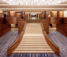 Un moderno vestíbulo de hotel con una escalera central, paneles de madera y una alfombra estampada.