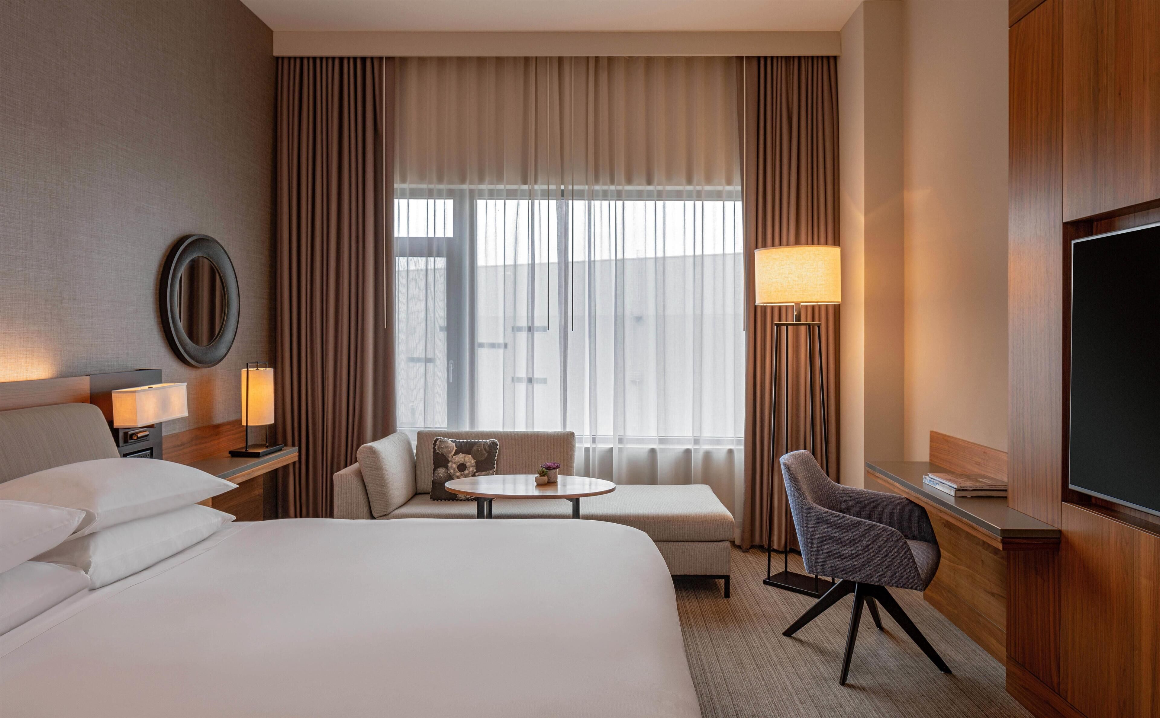 A hotel room with a bed, a sofa, a small table, a chair, a TV, and a window with curtains.
