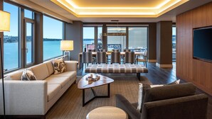 A modern hotel room with a sofa, armchair, round table, and a dining area with chairs.