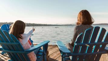 Two people sitting on blue Adirondack chairs facing a body of water with a forested shoreline.