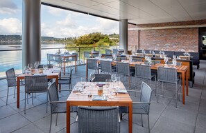 Enjoy a meal with a view of the serene lake from our outdoor dining area, featuring comfortable seating and a brick wall with large windows.