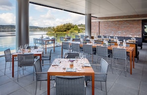 Enjoy a meal with a view of the serene lake from our outdoor dining area, featuring comfortable seating and a brick wall with large windows.