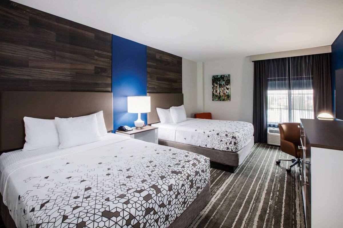 Habitación de hotel con dos camas, un escritorio, una silla y un ventanal con cortinas.