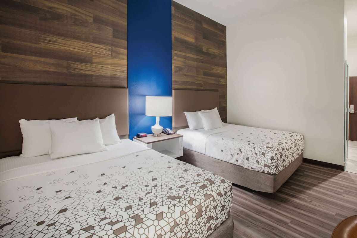 Habitación de hotel con dos camas, una mesita de noche con lámpara y una pared de acento de madera.