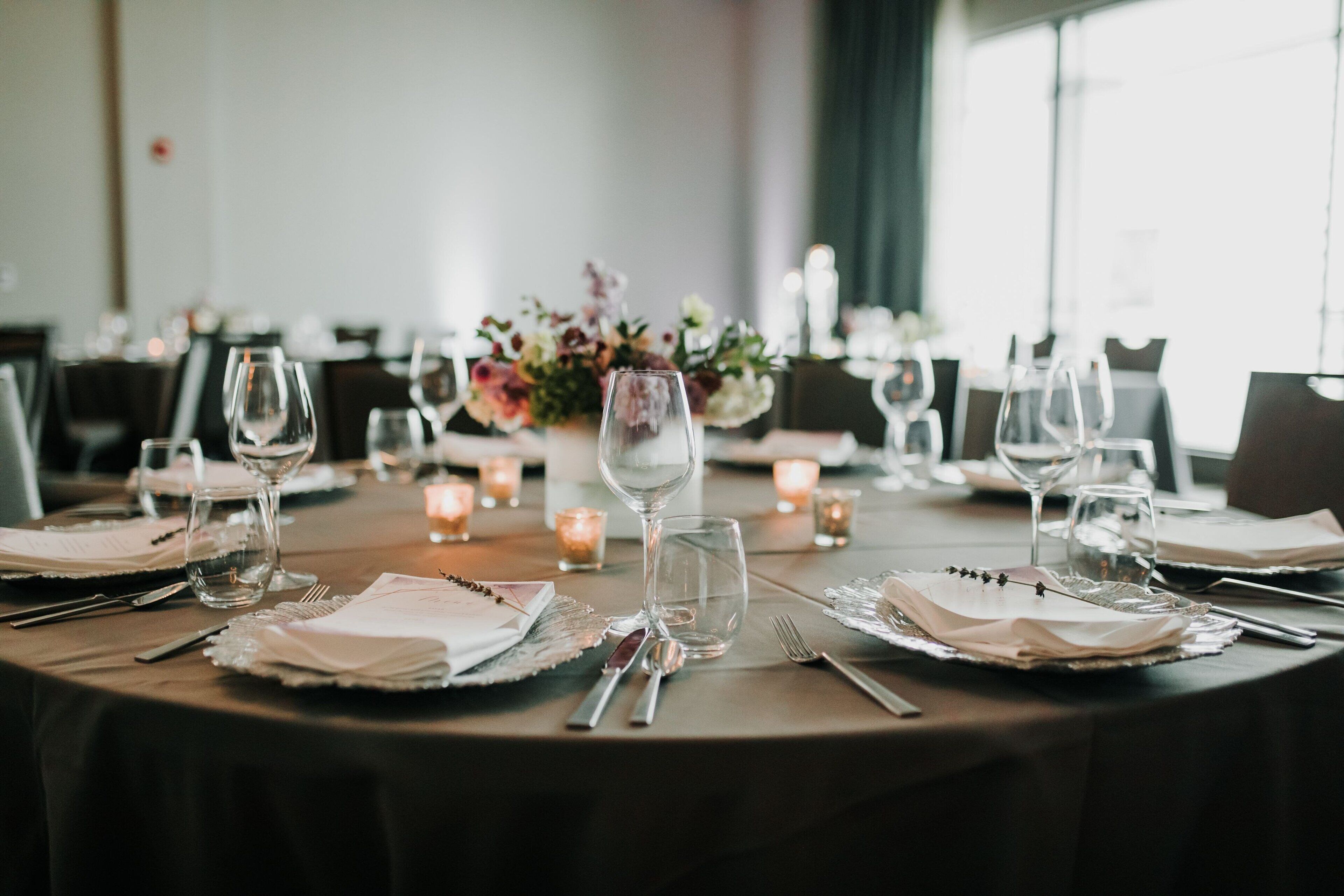 A dining table set with glassware, cutlery, and napkins, with candles and flowers in the background.