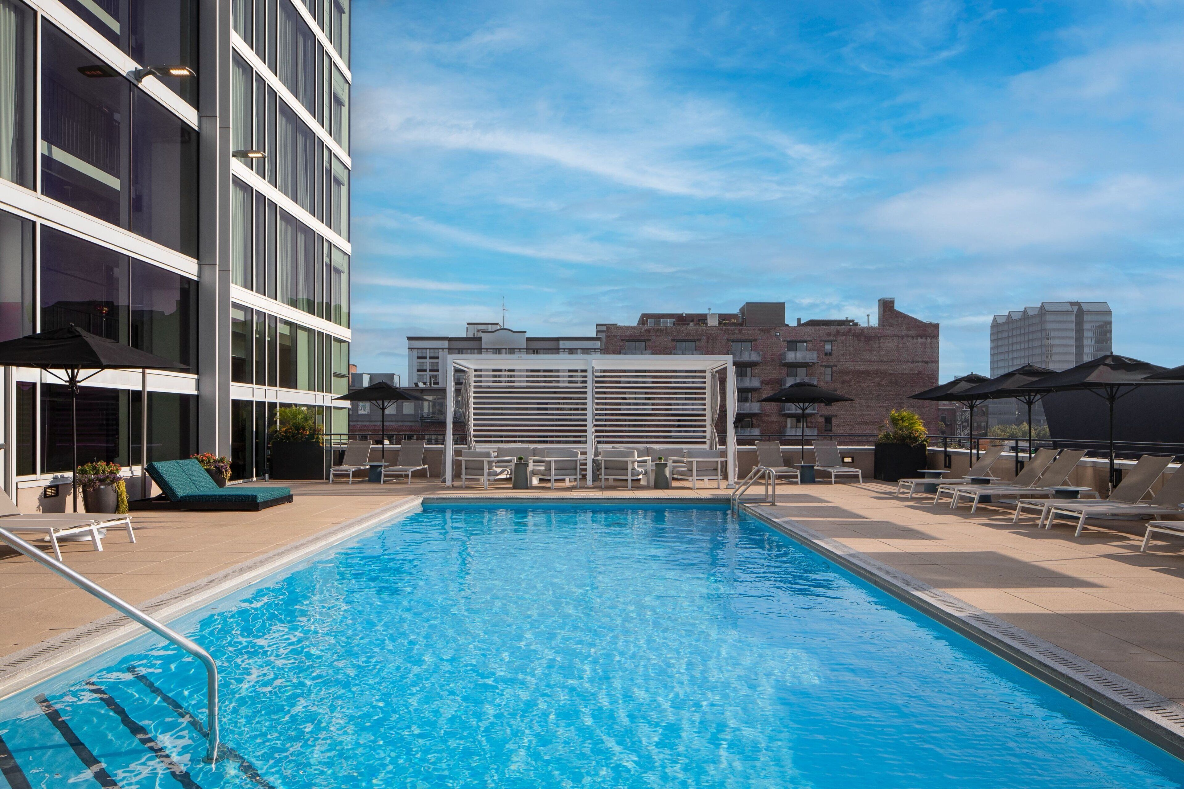 A rooftop pool with lounge chairs and umbrellas, surrounded by modern buildings.