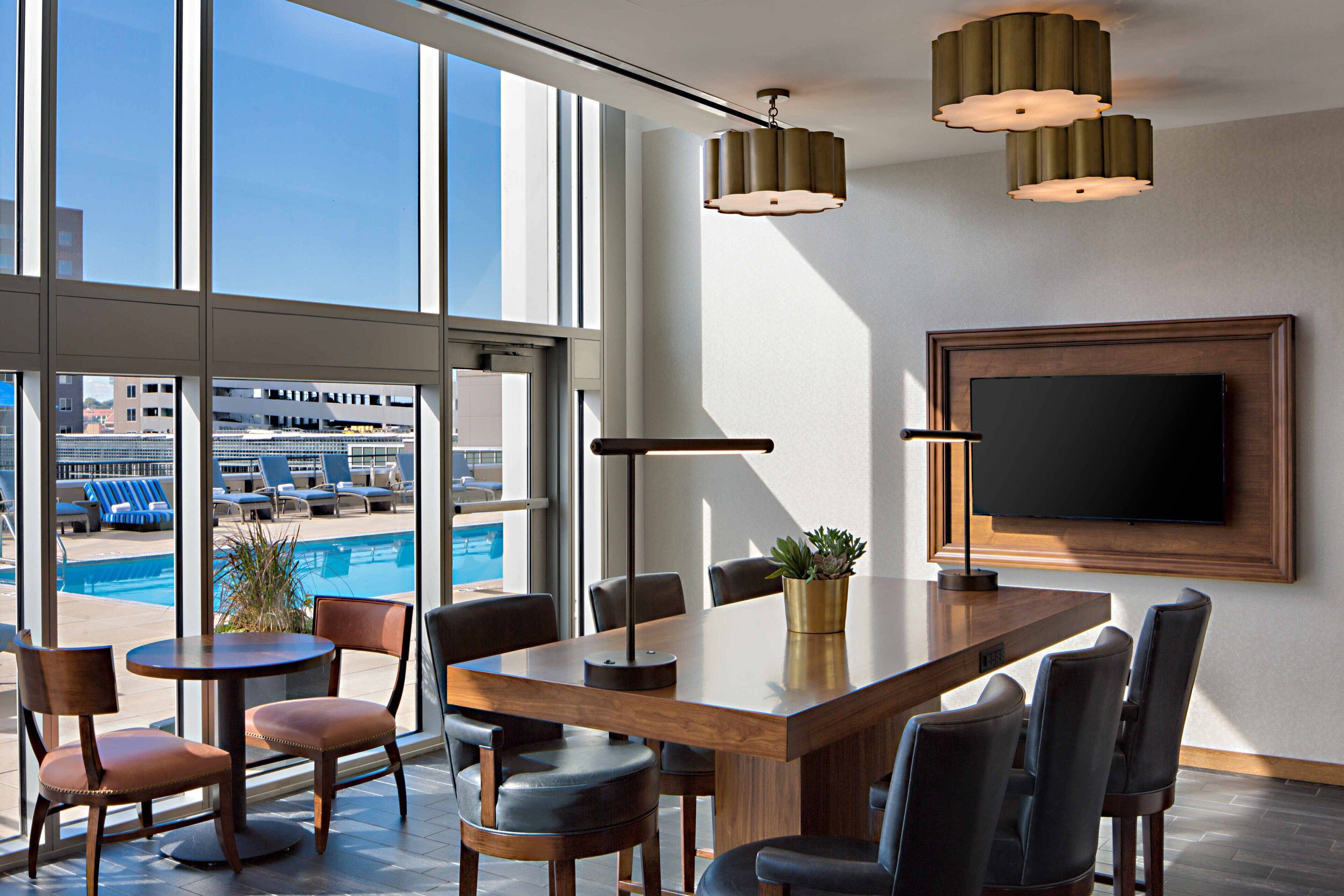 Una camera d'hotel moderna con una grande finestra vista piscina, un tavolo da pranzo in legno con sedie e un televisore a schermo piatto montato a parete.