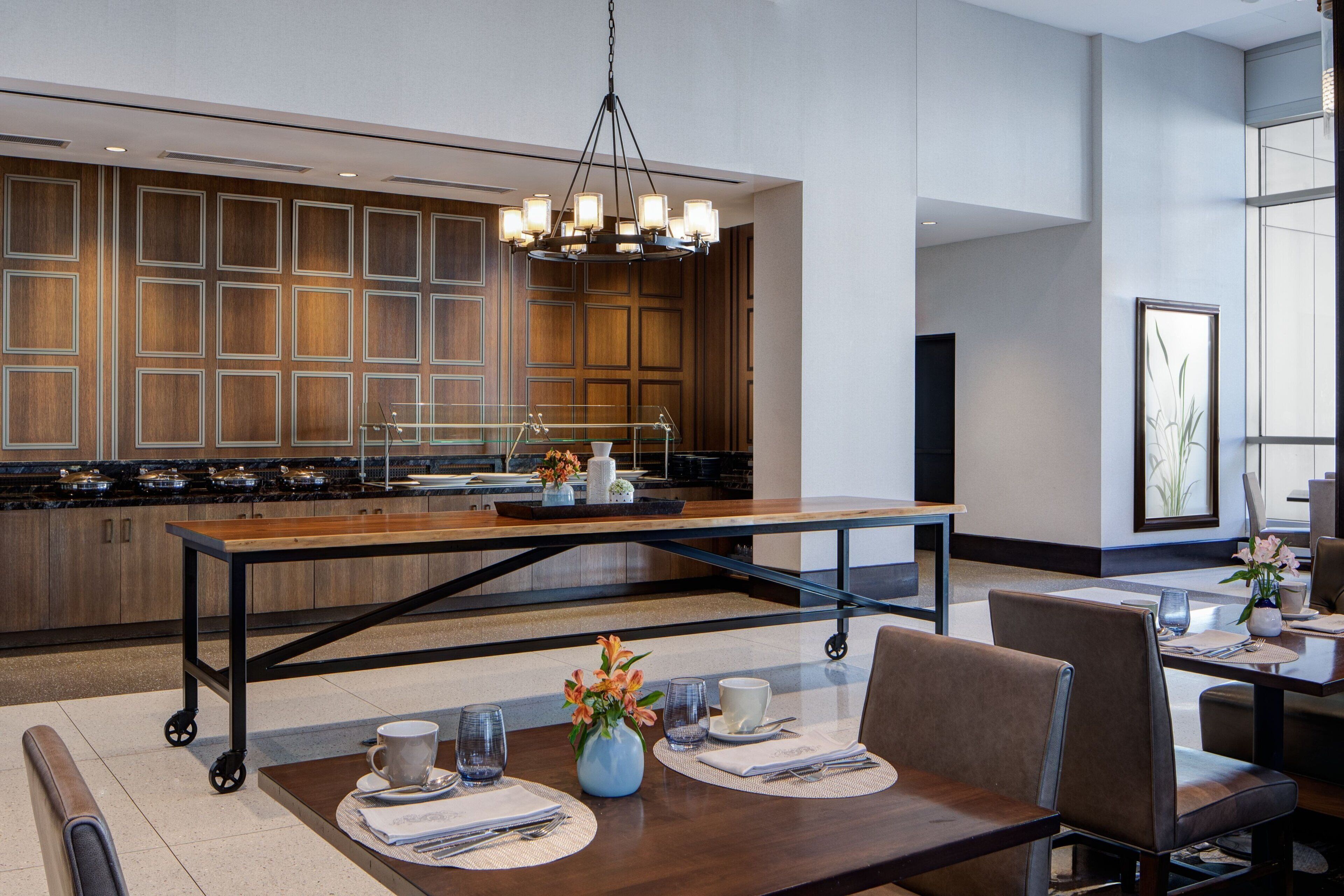 Uma cozinha moderna com uma ilha central, uma área de jantar com mesa posta para dois e uma grande janela que proporciona iluminação natural.