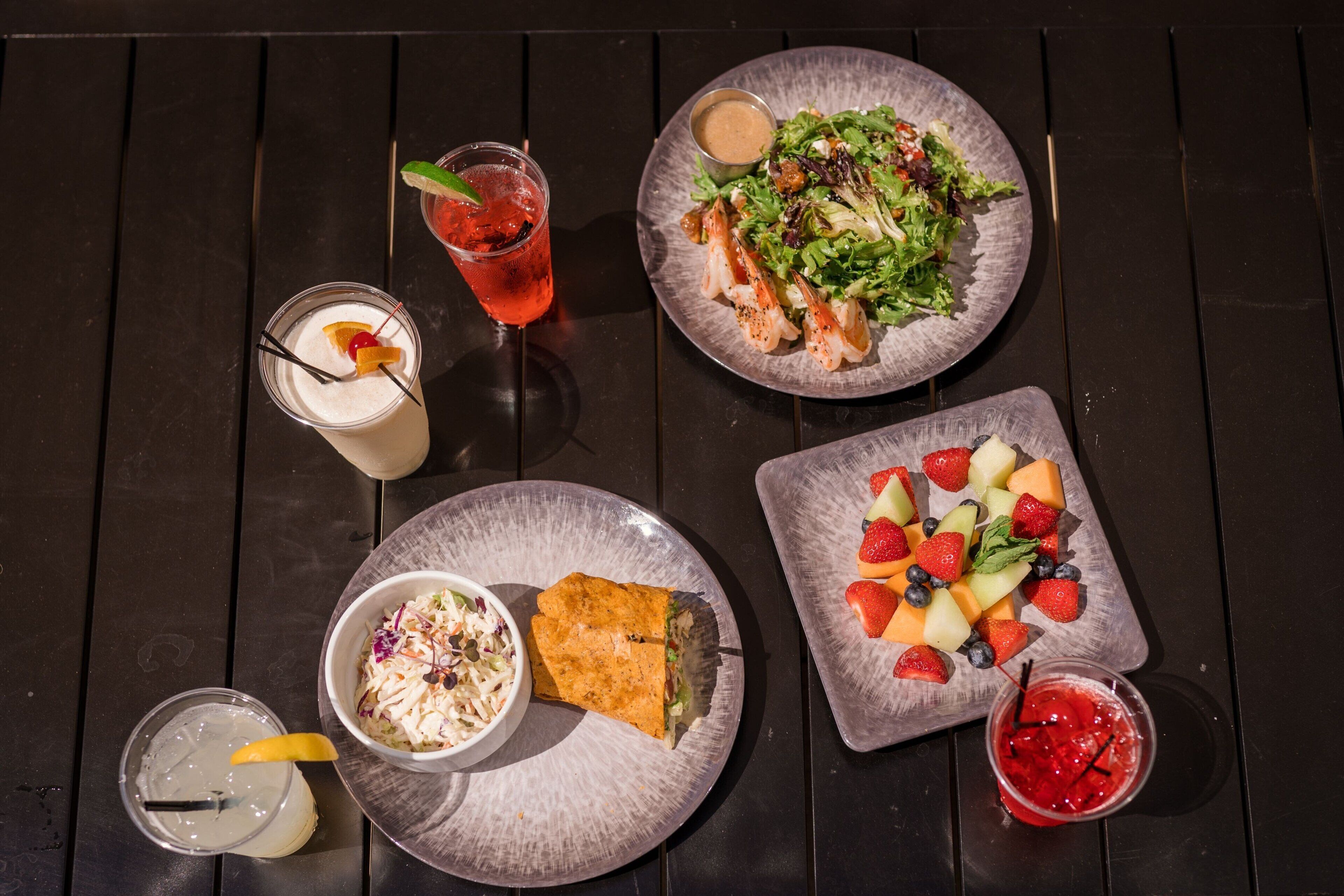 Een tafel met diverse etenswaren, waaronder een salade, een belegd broodje, een fruitplateau en drankjes.