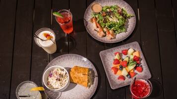 Een tafel met diverse etenswaren, waaronder een salade, een belegd broodje, een fruitplateau en drankjes.