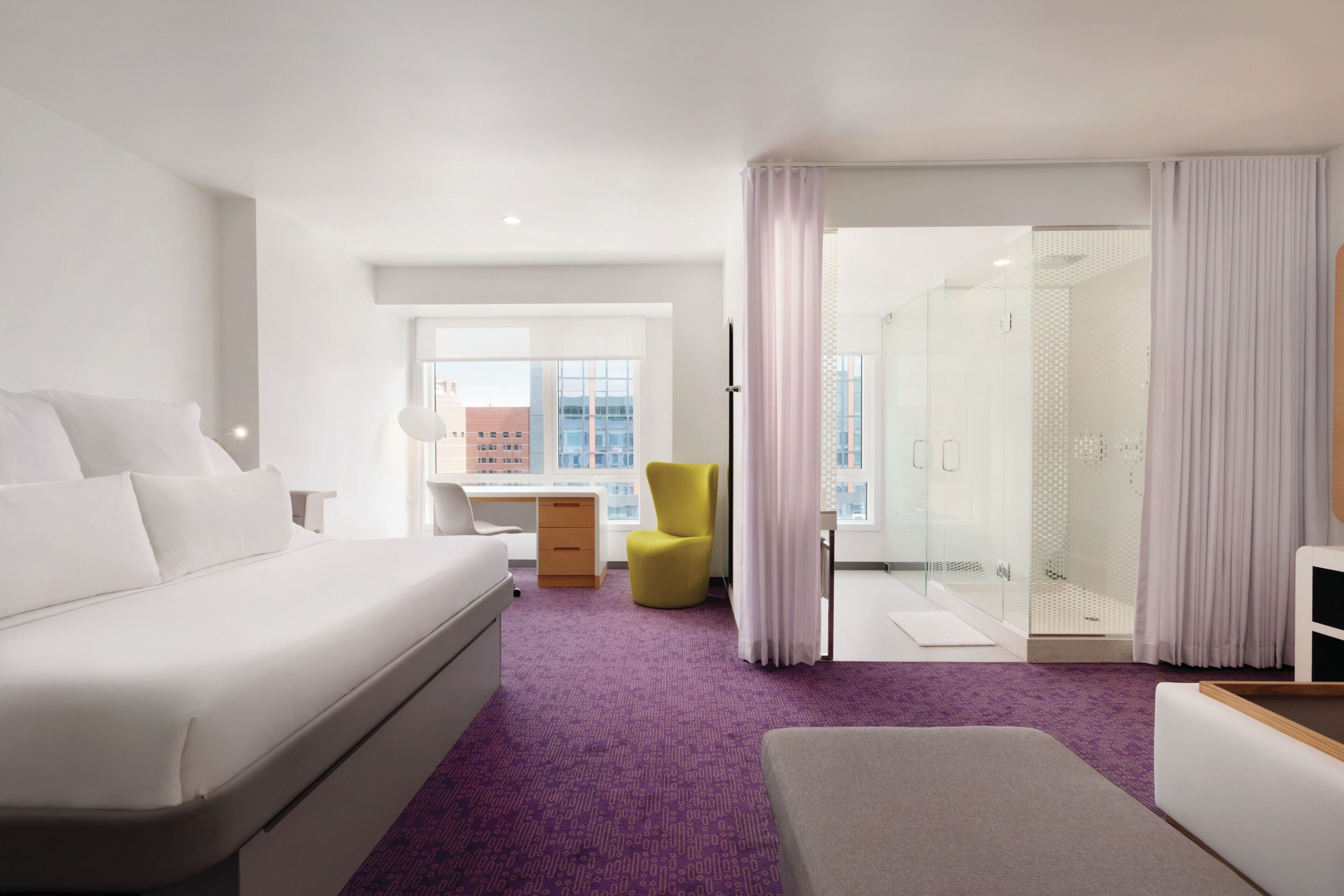 A hotel room with a bed, a desk, a chair, and a bathroom with a glass door.