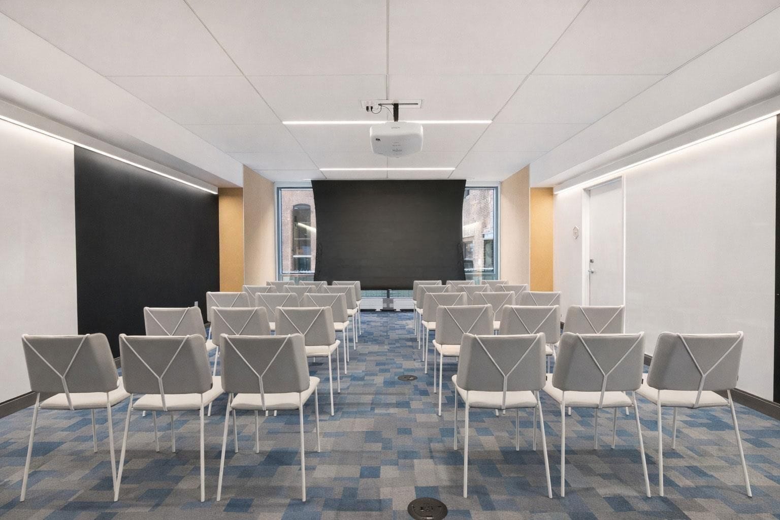 A conference room with rows of chairs facing a large screen, a projector, and a window with blinds.