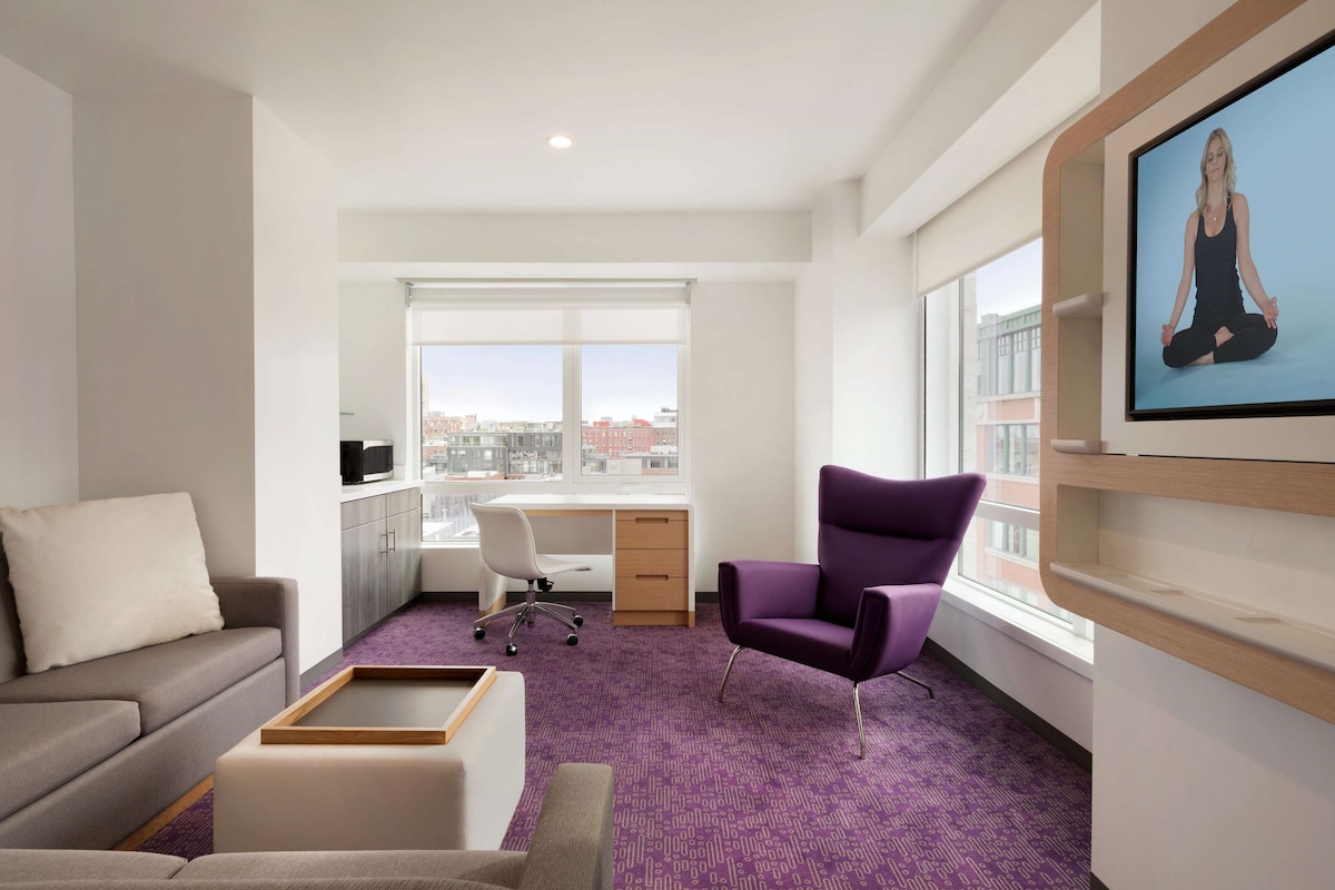 Ein modernes Zimmer mit einer Couch, einem Schreibtisch, einem violetten Sessel und einem großen Fenster mit Blick auf die Stadt.