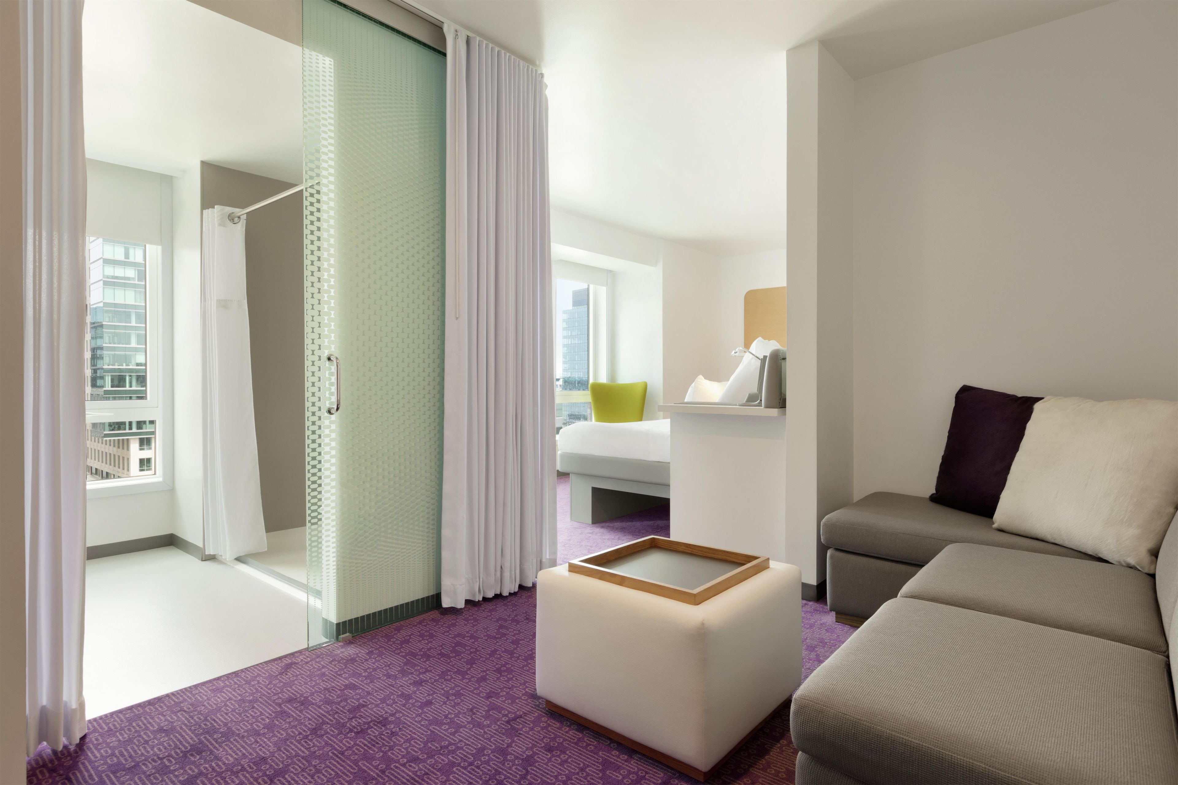 Una habitación de hotel moderna con una cama, un sofá, una mesita y vistas a un paisaje urbano.