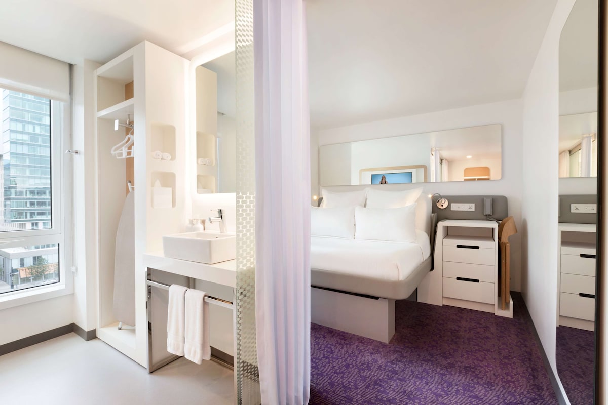 Ein modernes Hotelzimmer mit einem großen Bett, einem separaten Badezimmer, einem Schminktisch mit Spiegel und Blick auf die Stadt durch das Fenster.