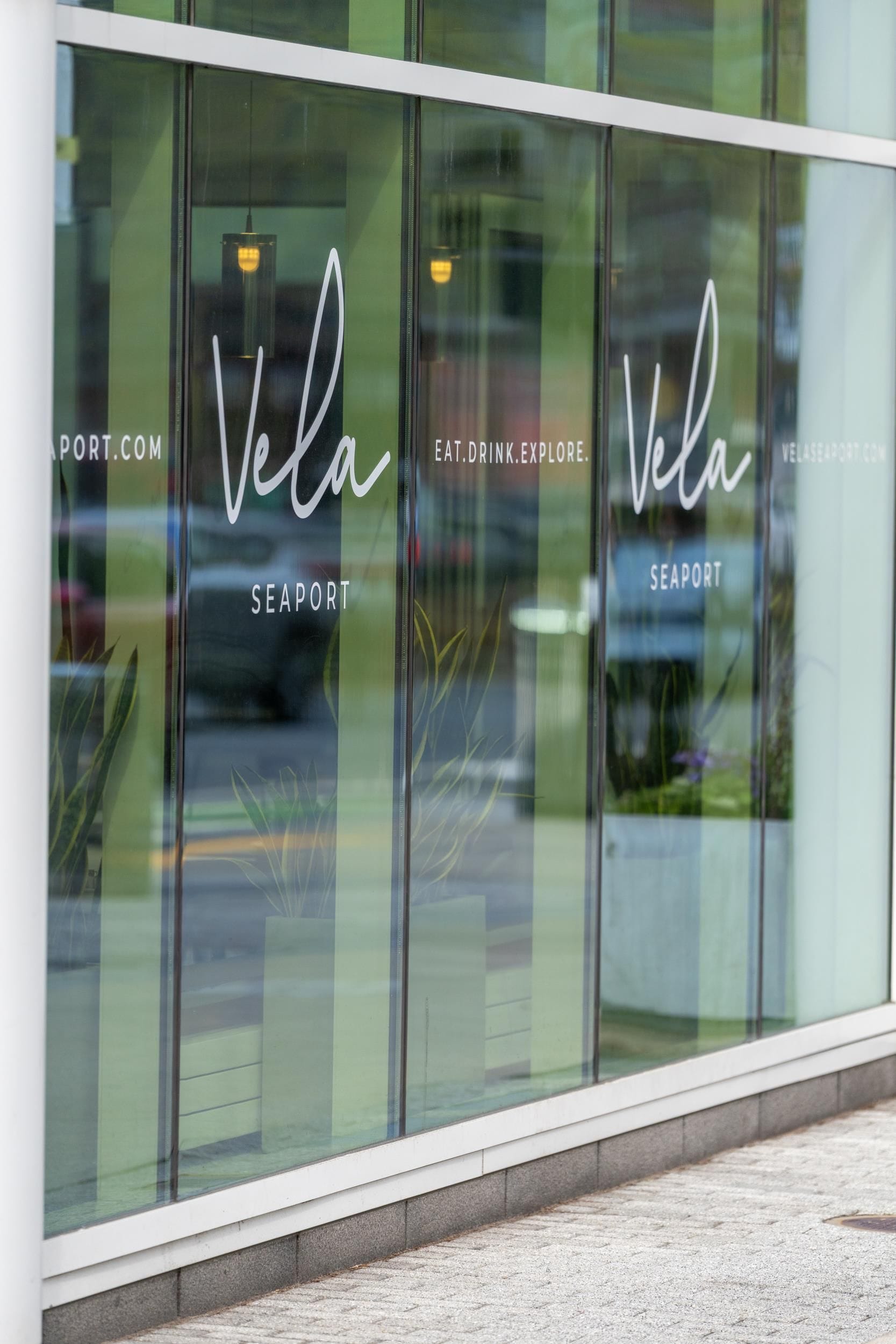 A storefront with the name 'Vela Seaport' and the website 'eat.drink.explore.com' displayed on the windows.