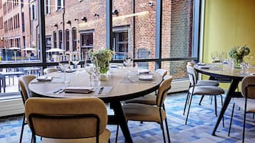 A restaurant with round tables, chairs, and a view of a brick building through large windows.