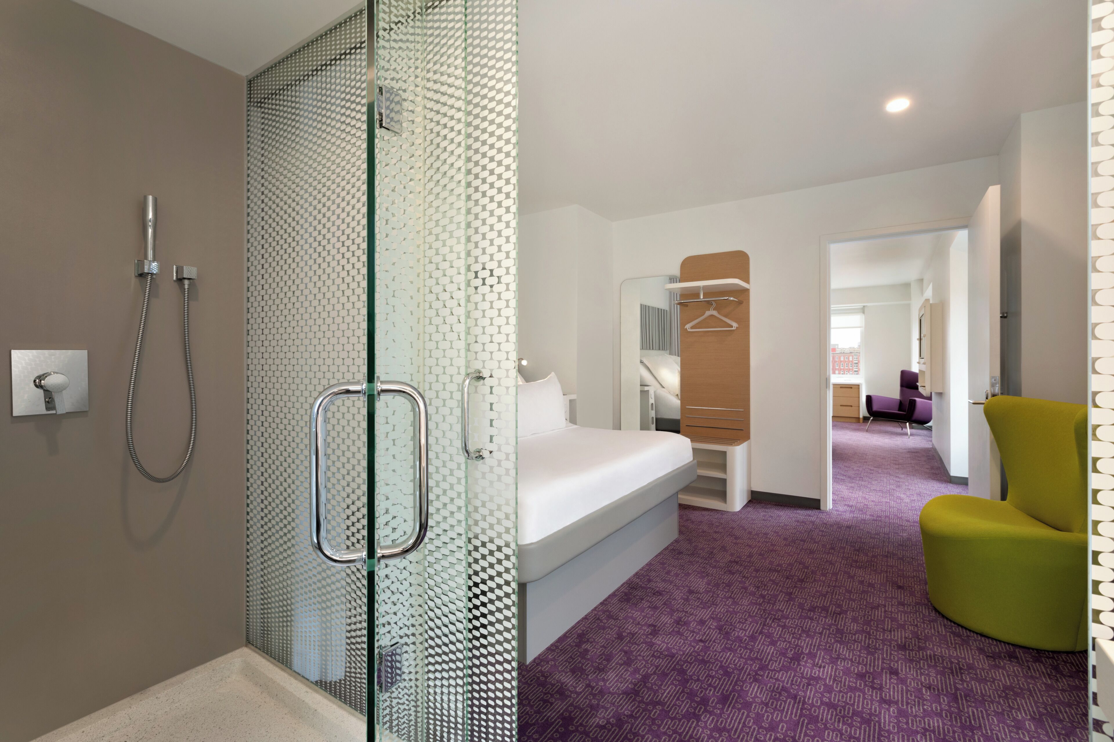Habitación de hotel con ducha, cama, una silla y un pasillo que conduce a otra habitación.