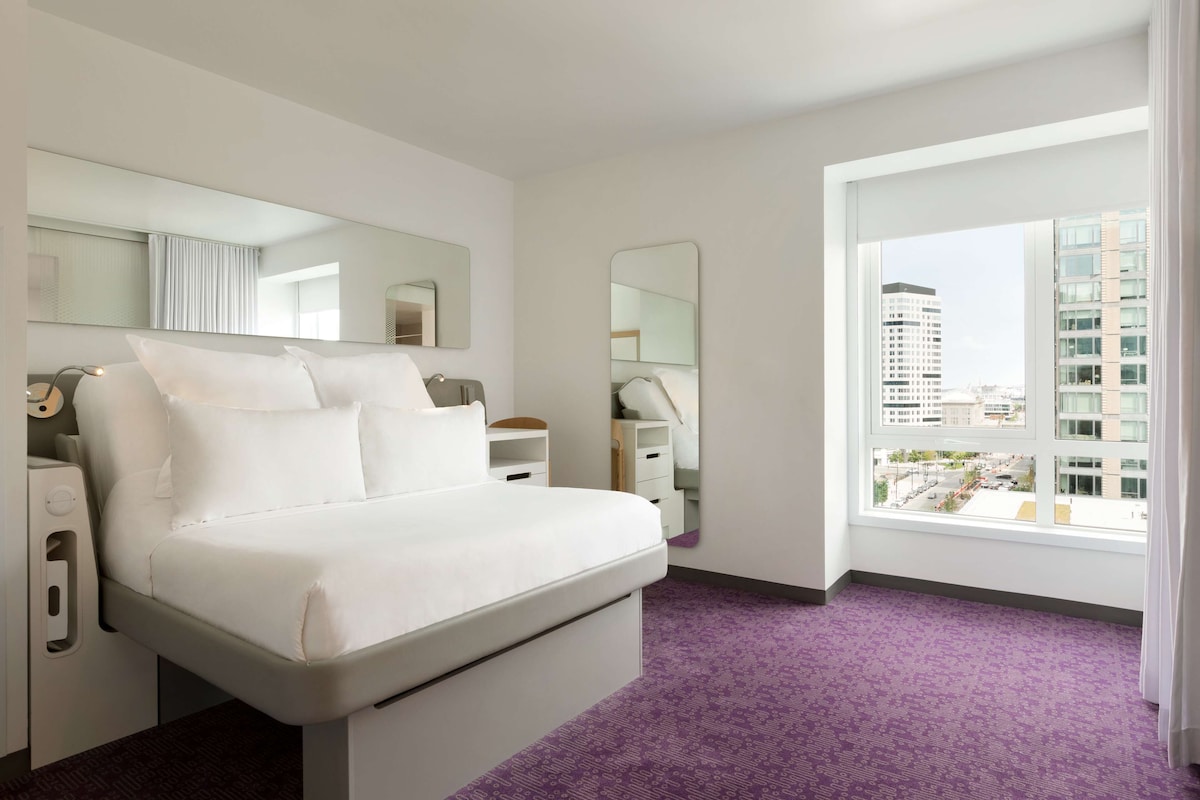 Ein modernes Hotelzimmer mit einem großen Bett, einem Schminktisch, einem Spiegel und einem Fenster mit Blick auf die Stadt.