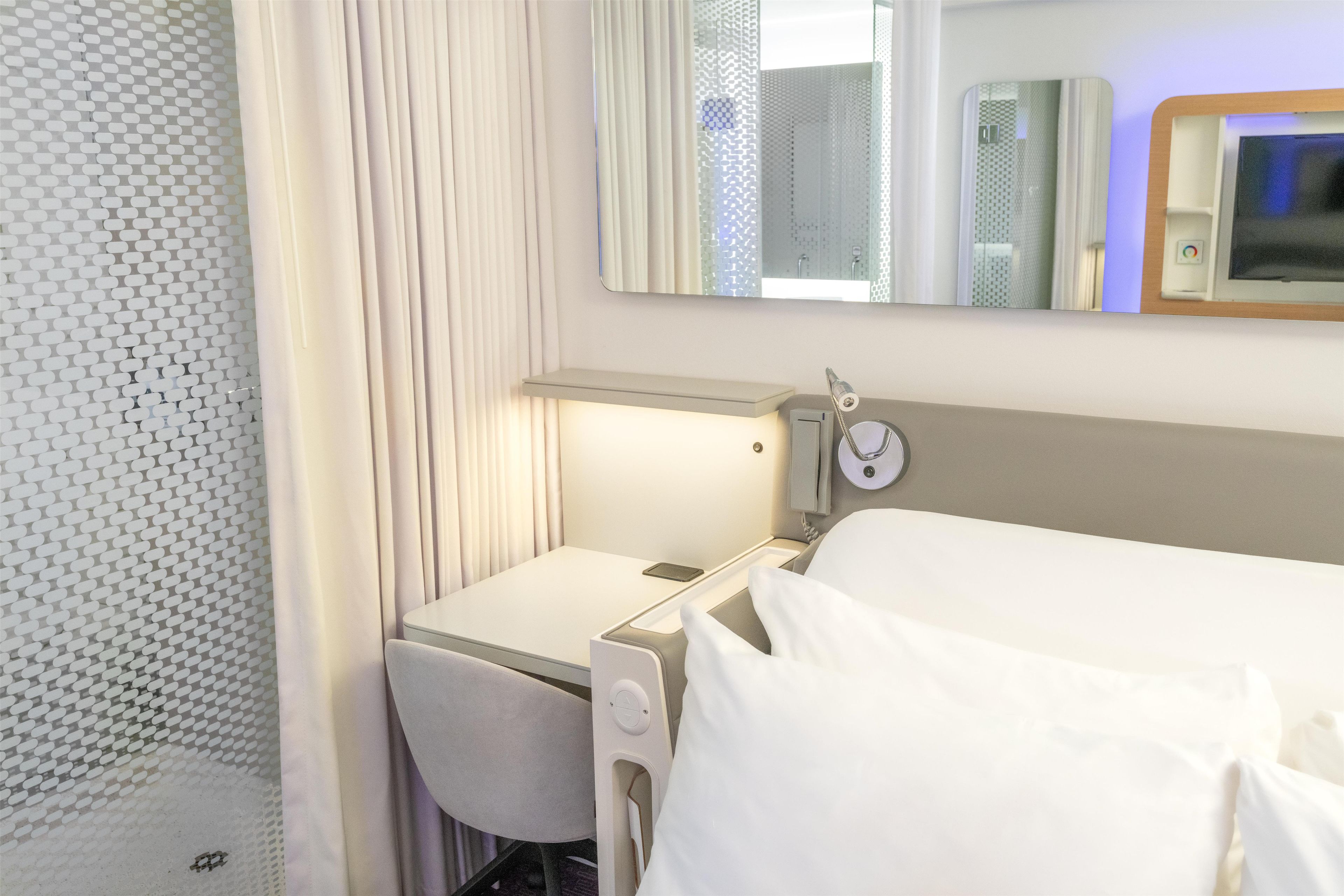 A hotel room with a bed, a bedside table, a mirror, and a window with curtains.