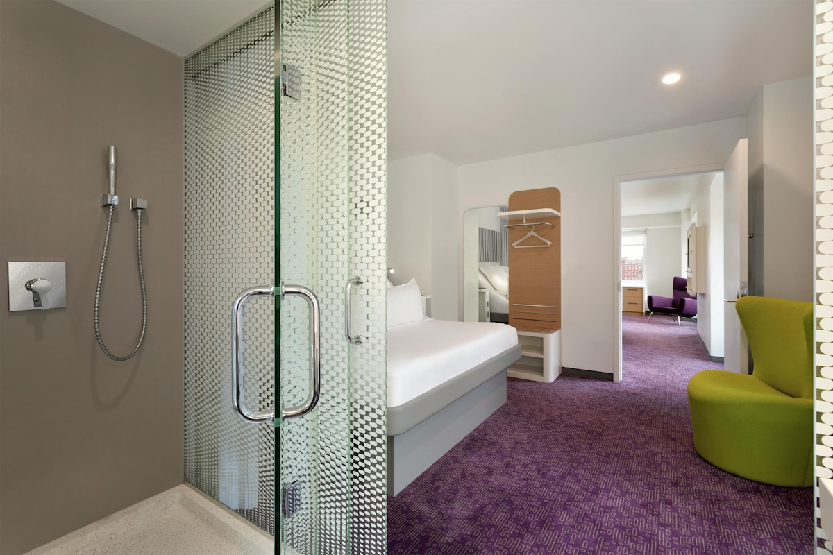 A hotel room with a shower, a bed, a chair, and a hallway leading to another room.