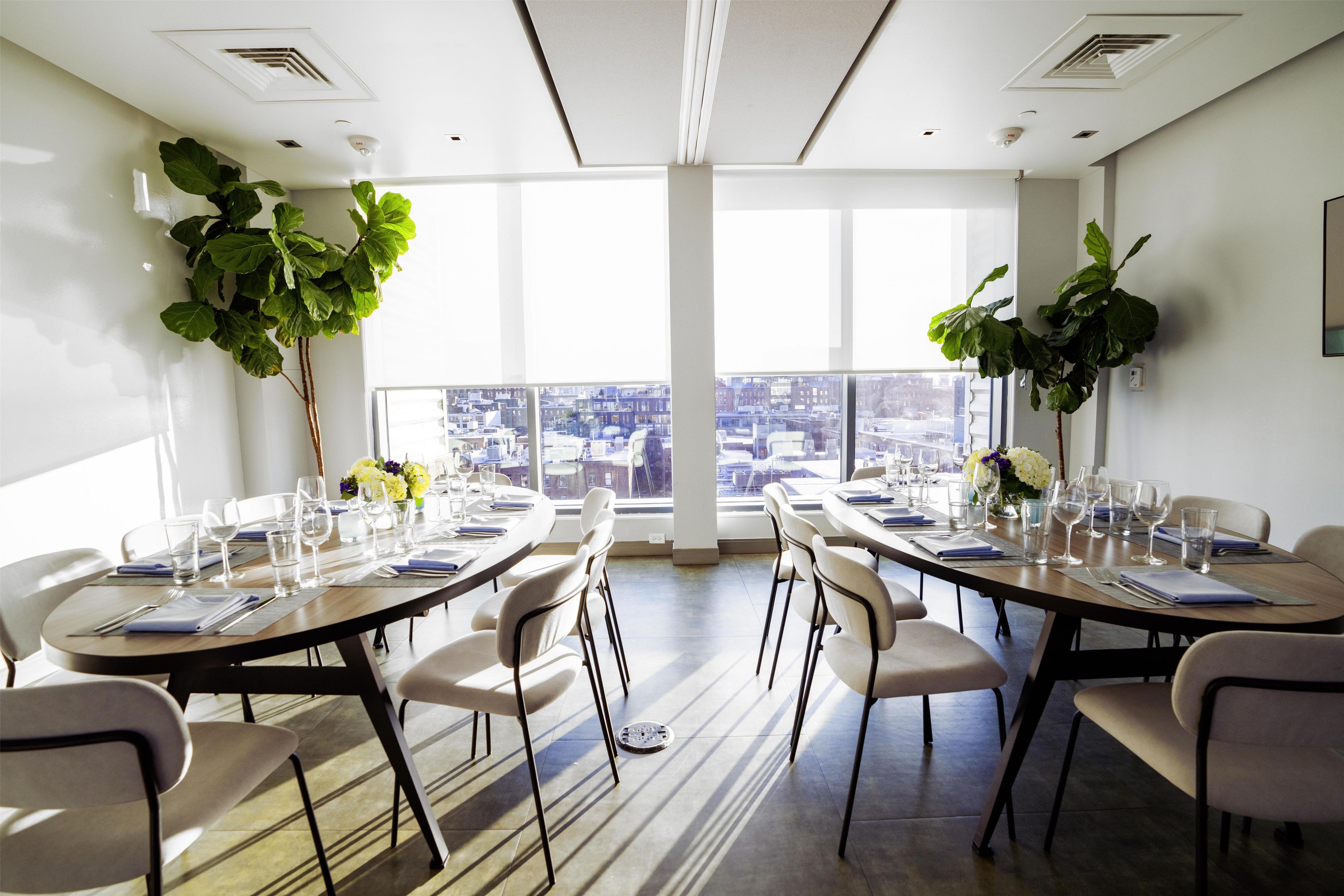 A conference room with a panoramic city view, equipped with tables, chairs, and a large potted plant.