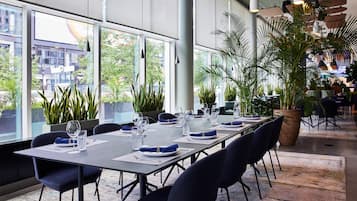Un comedor moderno con una mesa larga dispuesta para una comida, rodeado de ventanas amplias y plantas de interior.