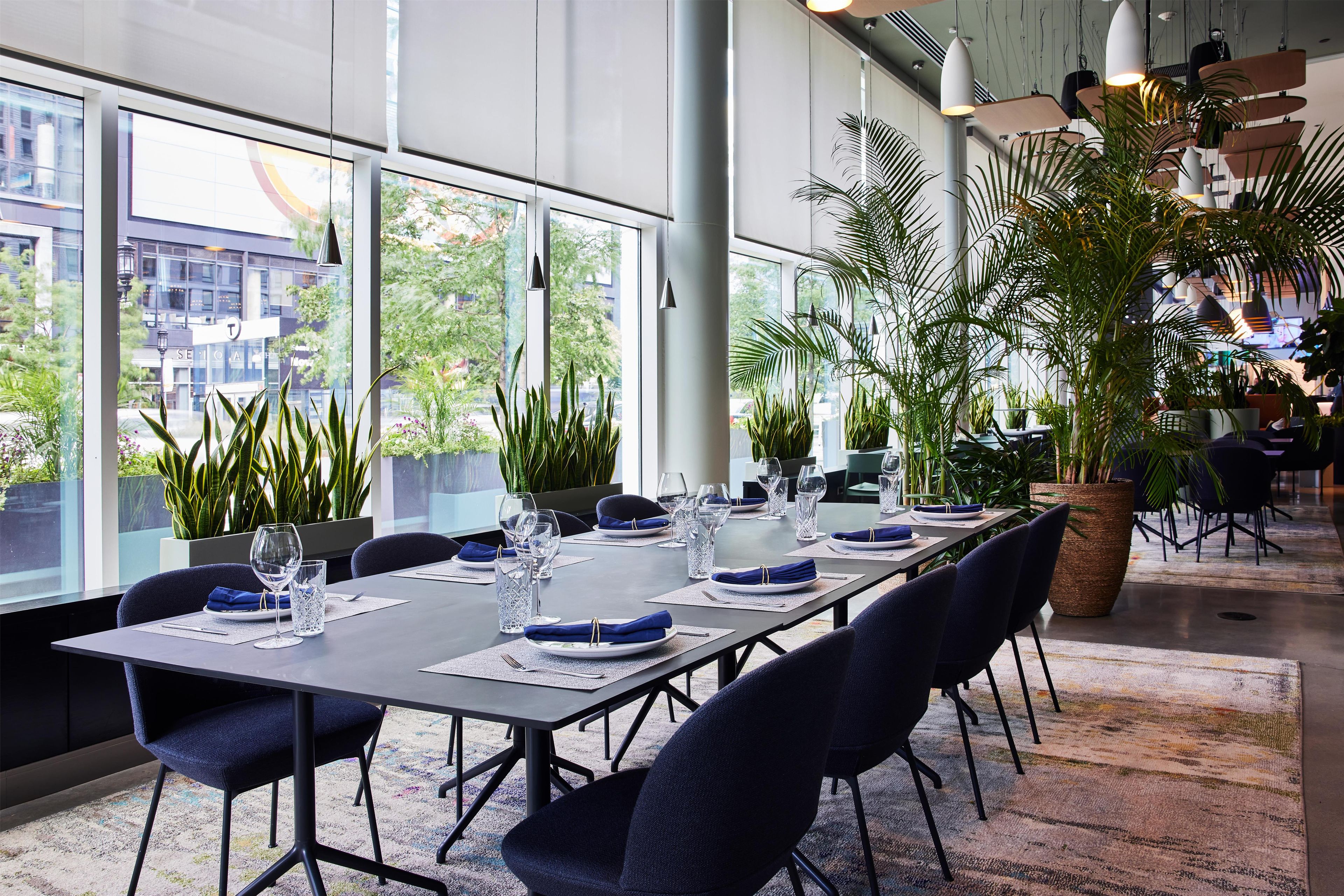 A modern dining area with a long table set for a meal, surrounded by large windows and indoor plants.