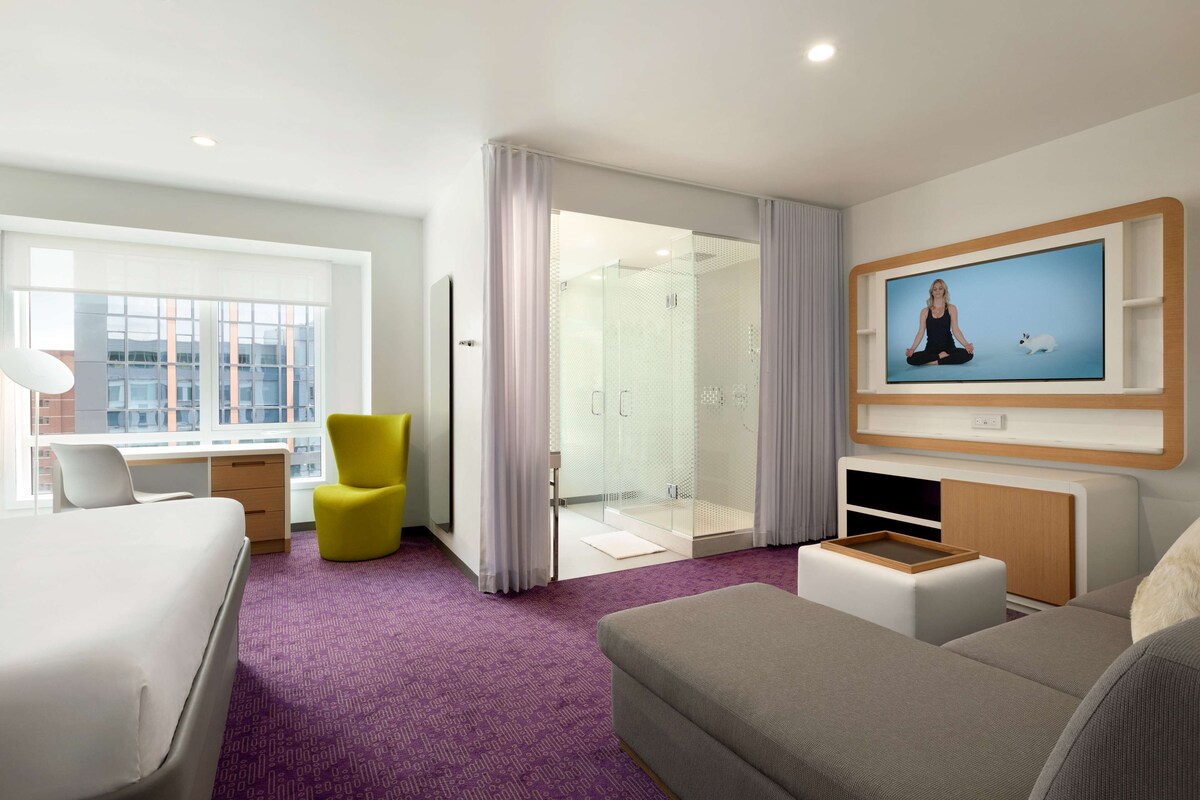 Ein modernes Hotelzimmer mit Bett, Schreibtisch, Stuhl, Fernseher und Duschbereich.