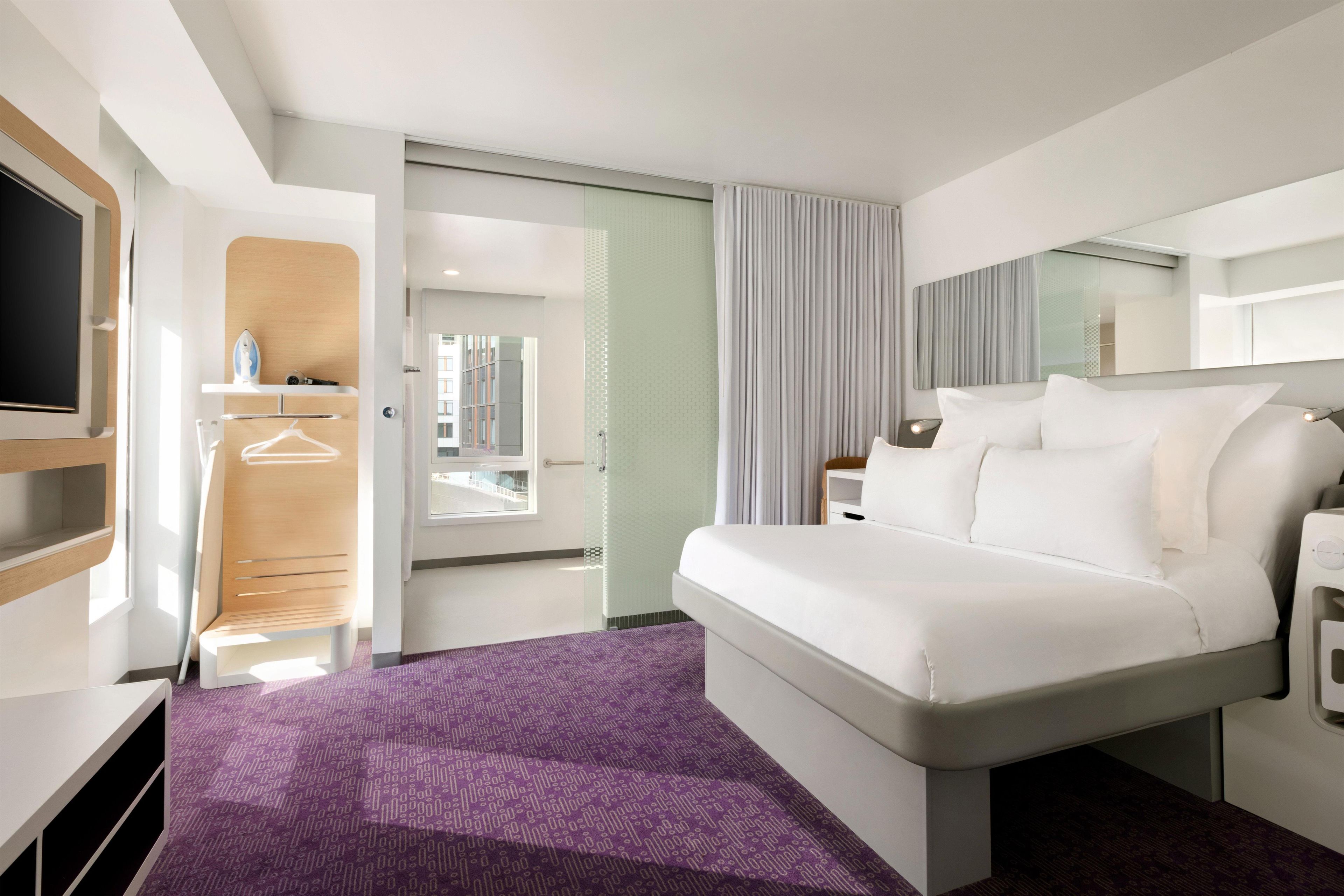 A modern hotel room with a large bed, a TV, a wardrobe, and a bathroom visible through an open door.