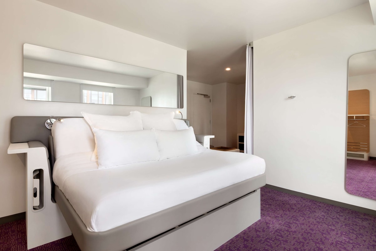 Ein modernes Hotelzimmer mit einem großen Bett, einem Nachttisch, einem Spiegel und einem violetten Teppich.