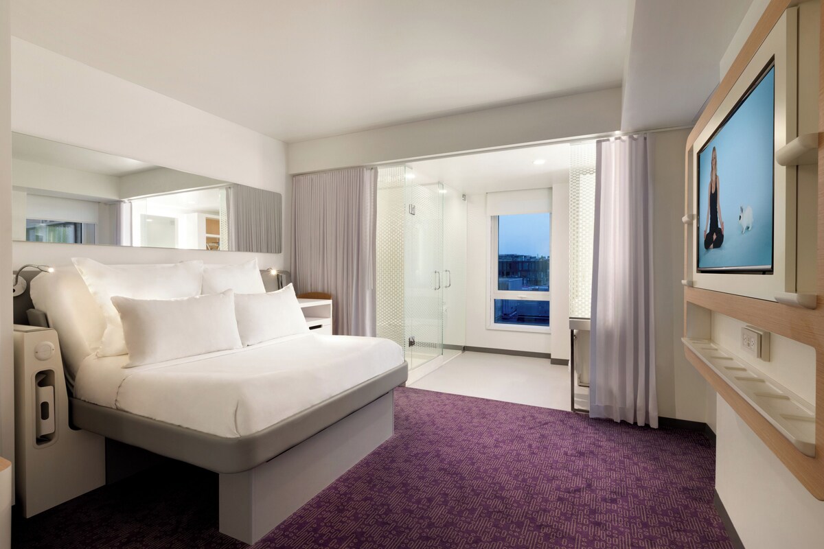 A modern hotel room with a large bed, a bathroom with a glass shower, and a view of the city.