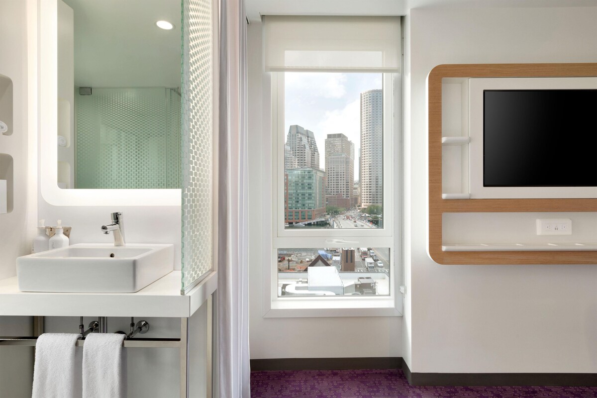 A modern bathroom with a large mirror, a sink, and a window offering a city view.