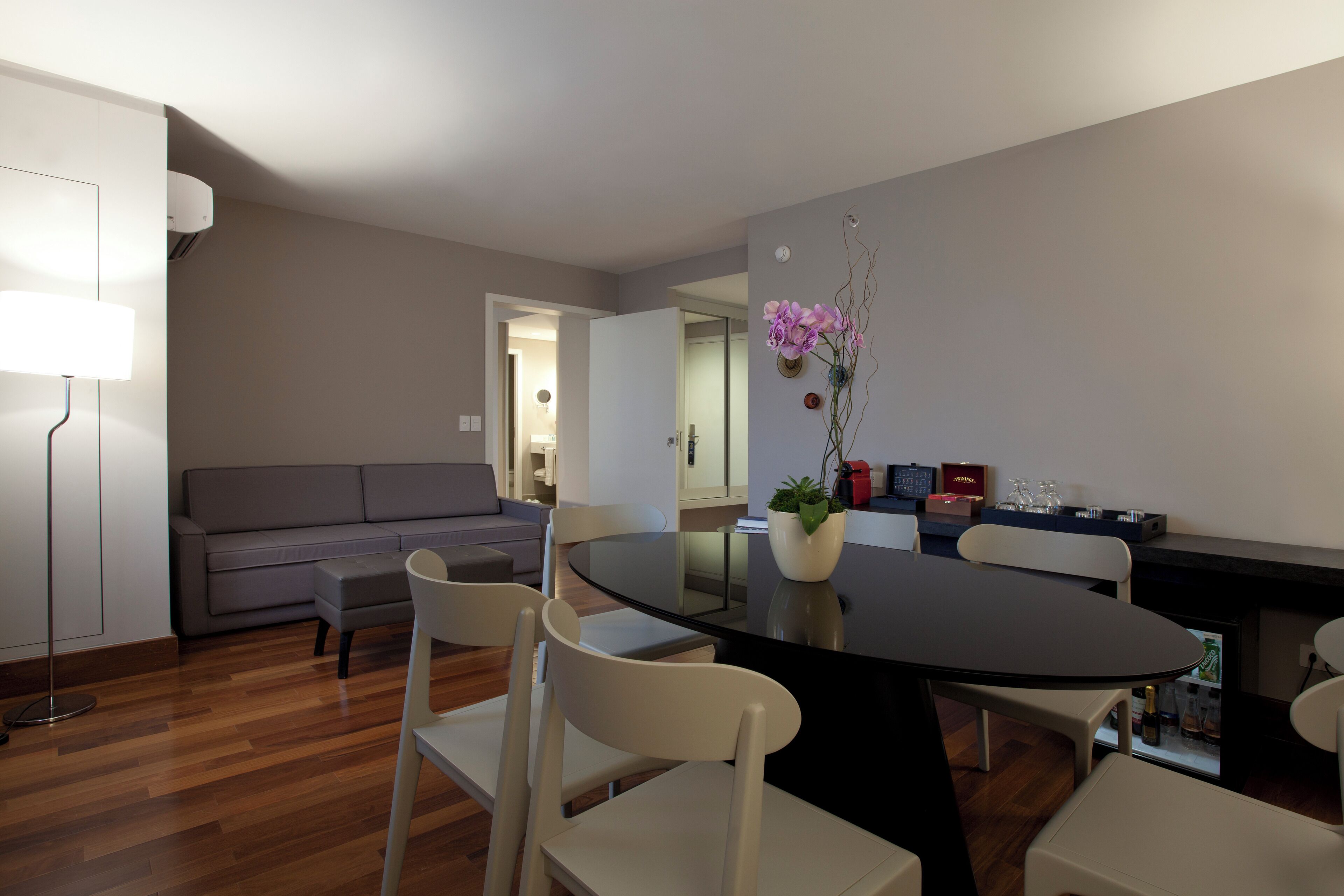 A modern dining area with a round table, white chairs, and a potted orchid. A grey sofa and a floor lamp are visible in the background.
