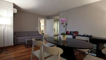A modern dining area with a round table, white chairs, and a potted orchid. A grey sofa and a floor lamp are visible in the background.